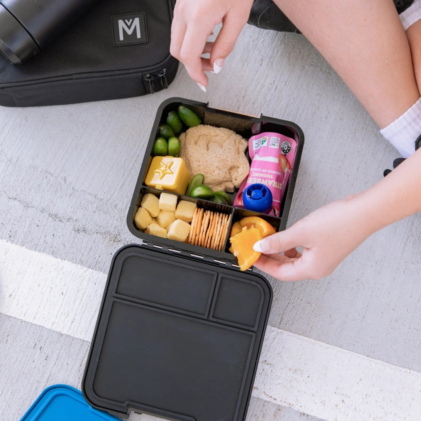 Birdseye view of a childs black bento lunchbox filled with food and hands reaching for an orange.