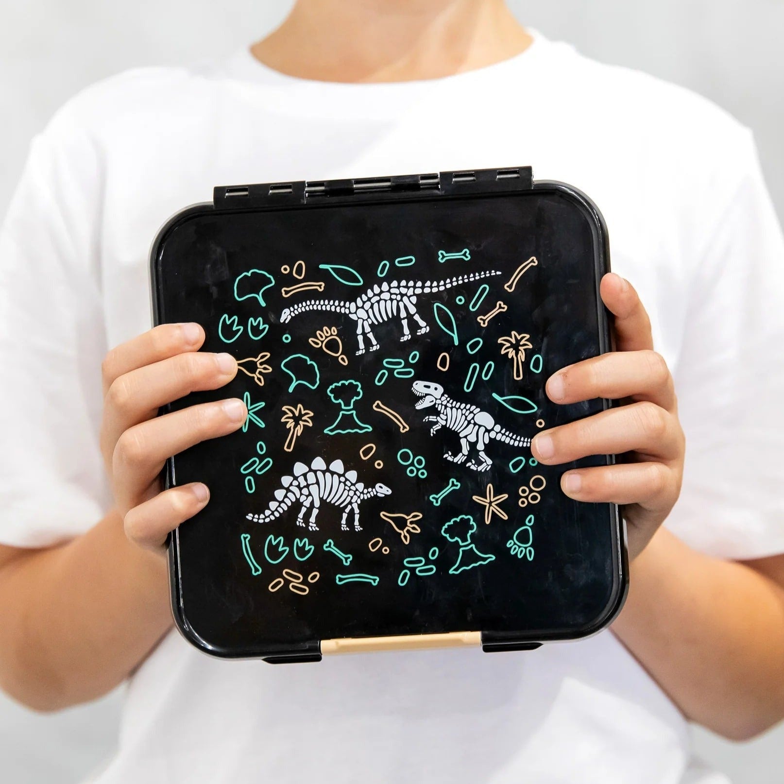 Close up of a childs hands holding a black lunchbox with green and white dinosaur print on the lid.