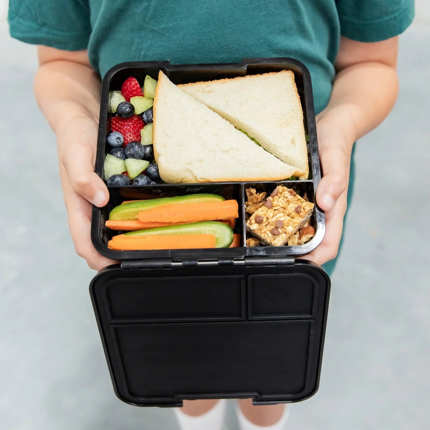 Childs hands holding a bento lunchbox in black filled with fruit, sandwiches and granola bars.