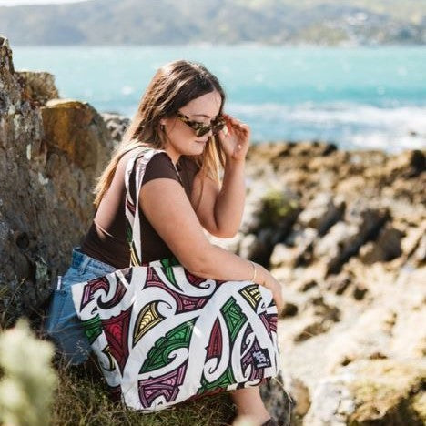 Woman sitting outdoors with a Tote bag with red, pink, green, yellow, white & black maori design.