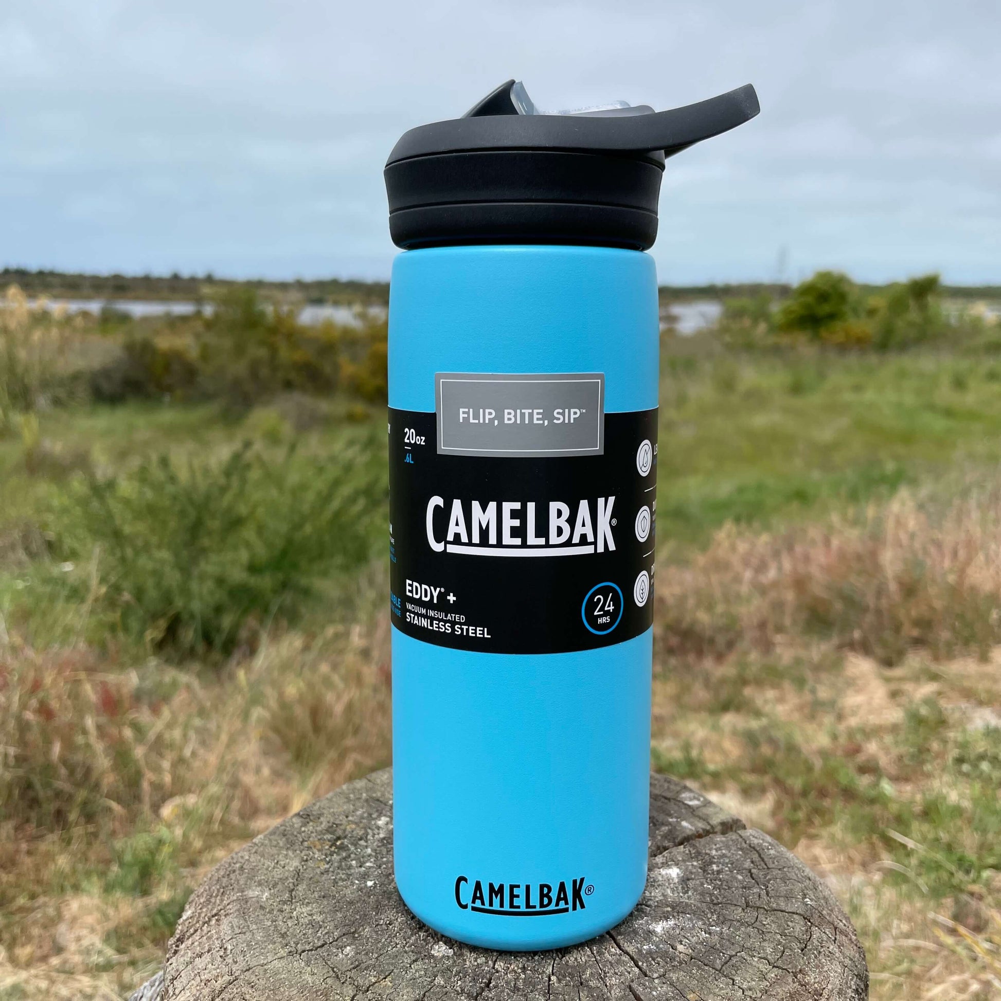 Stainless steel camelbak sipper bottle in Nordic Blue colour with black lid. Sitting on a wooden fence post with a blurry Estuary scene in the background.