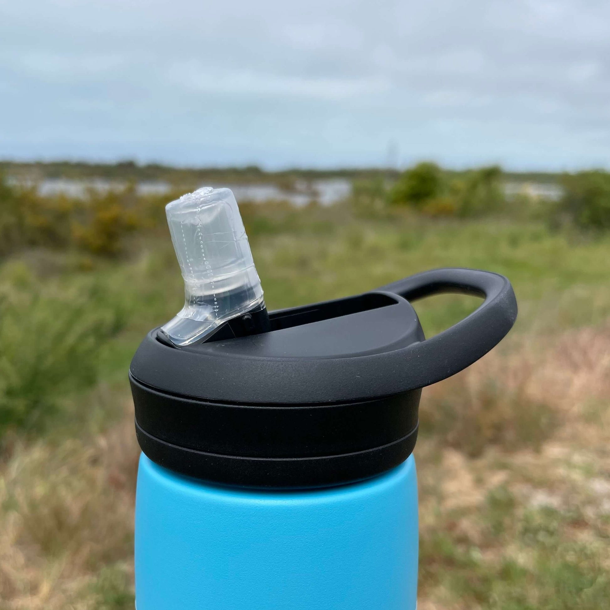 Close up of a black Camelbak lid with sipper top on a blue drink bottle. Blurred out estuary in the background. 