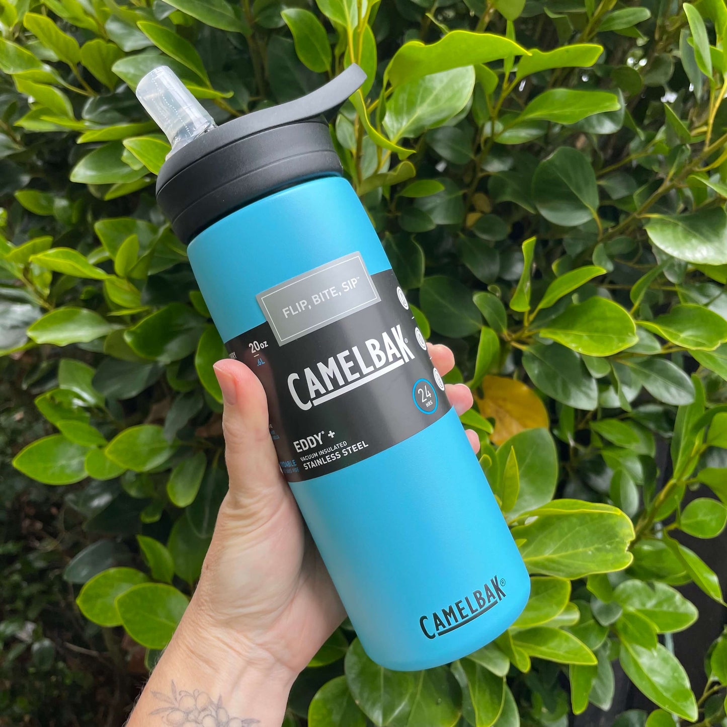 Persons hand holding a 600ml blue stainless camelbak drink bottle with sipper open in front of a green hedge.