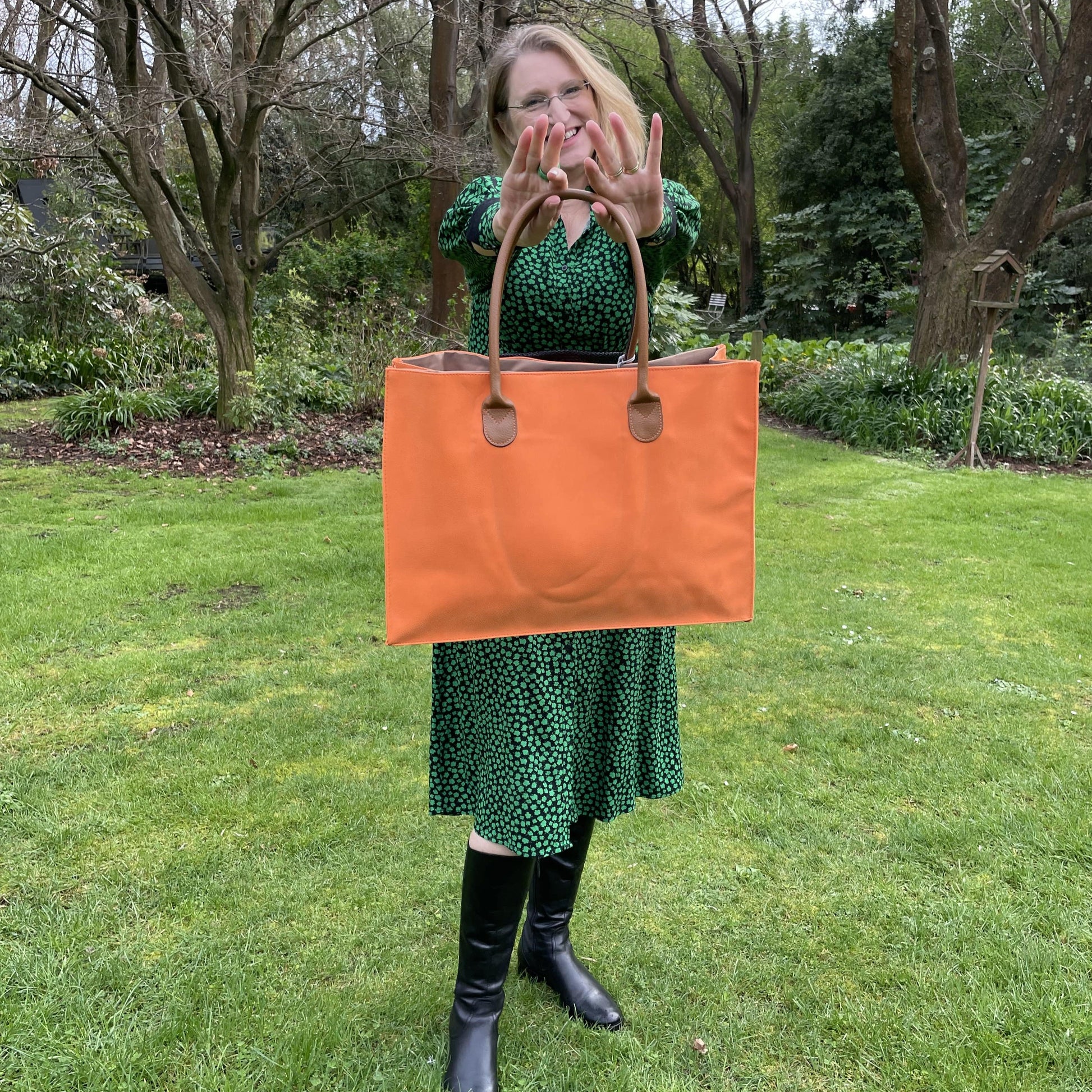 Blonde woman in a black and green dress standing in a garden setting, holding a large bright orange tote bag out in front of her.