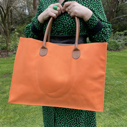 Close up of a bright orange tote bag with brown leather look handles, being held by a woman in a black and green dress in a garden setting.