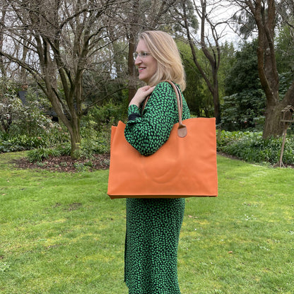 Blonde woman wearing a black and green dress standing outdoors in a garden with a bright orange tote bag over her shoulder.