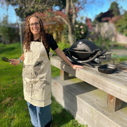 Woman wearing calico apron with black sketch of a Tui bird and kowhai flowers.