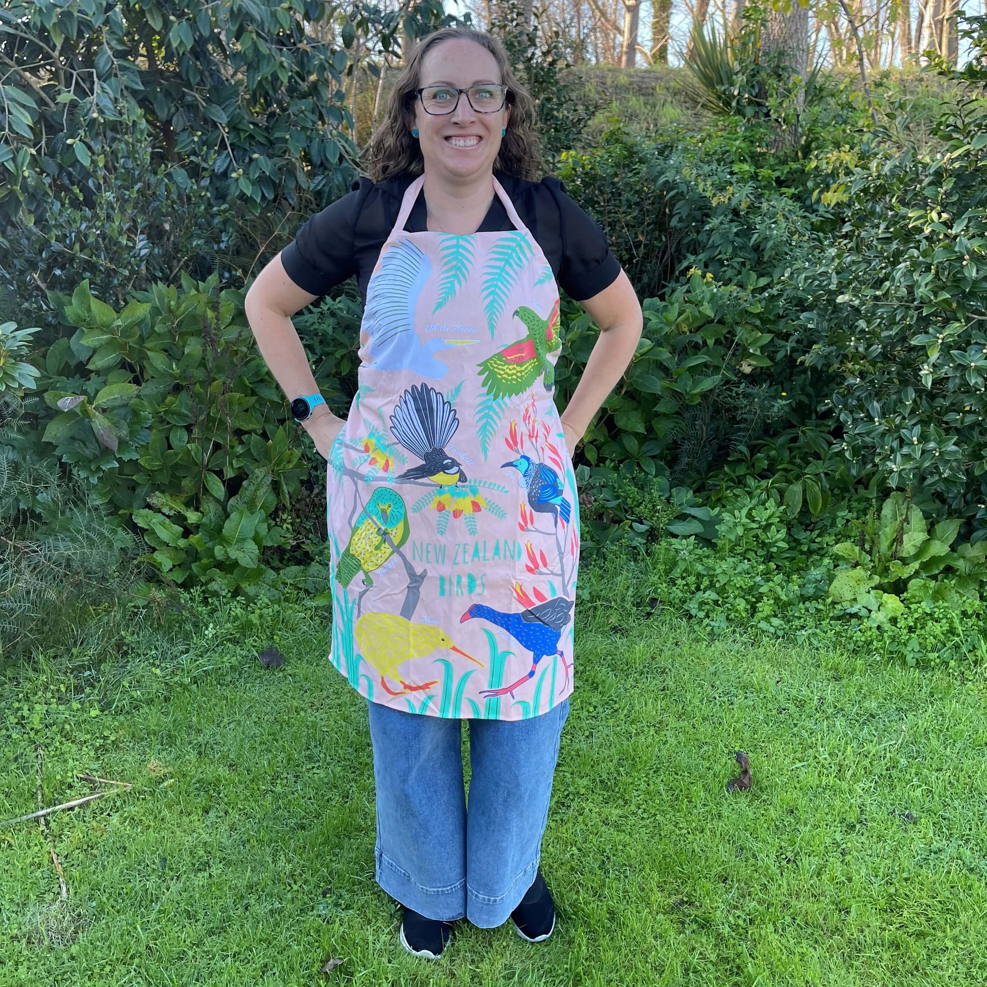 Woman wearing a peach coloured apron with bold and colourful NZ birds printed on it.