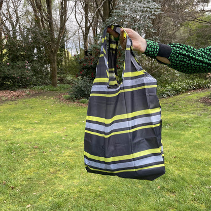 Womans hand holding a striped black, grey and lime green reusable shopping tote bag with a lush green garden in the background.