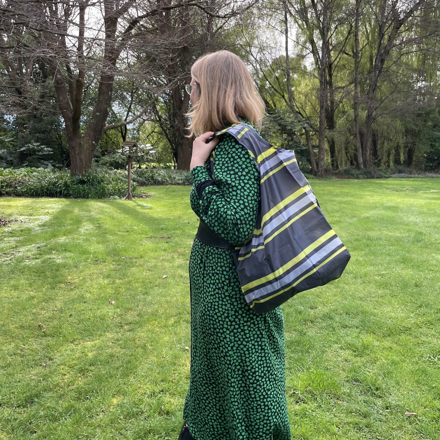 Woman standing in a garden in a black and green dress with her back to the camera. She has a bstriped black, grey and lime green reusable shopping tote bag over her shoulder.