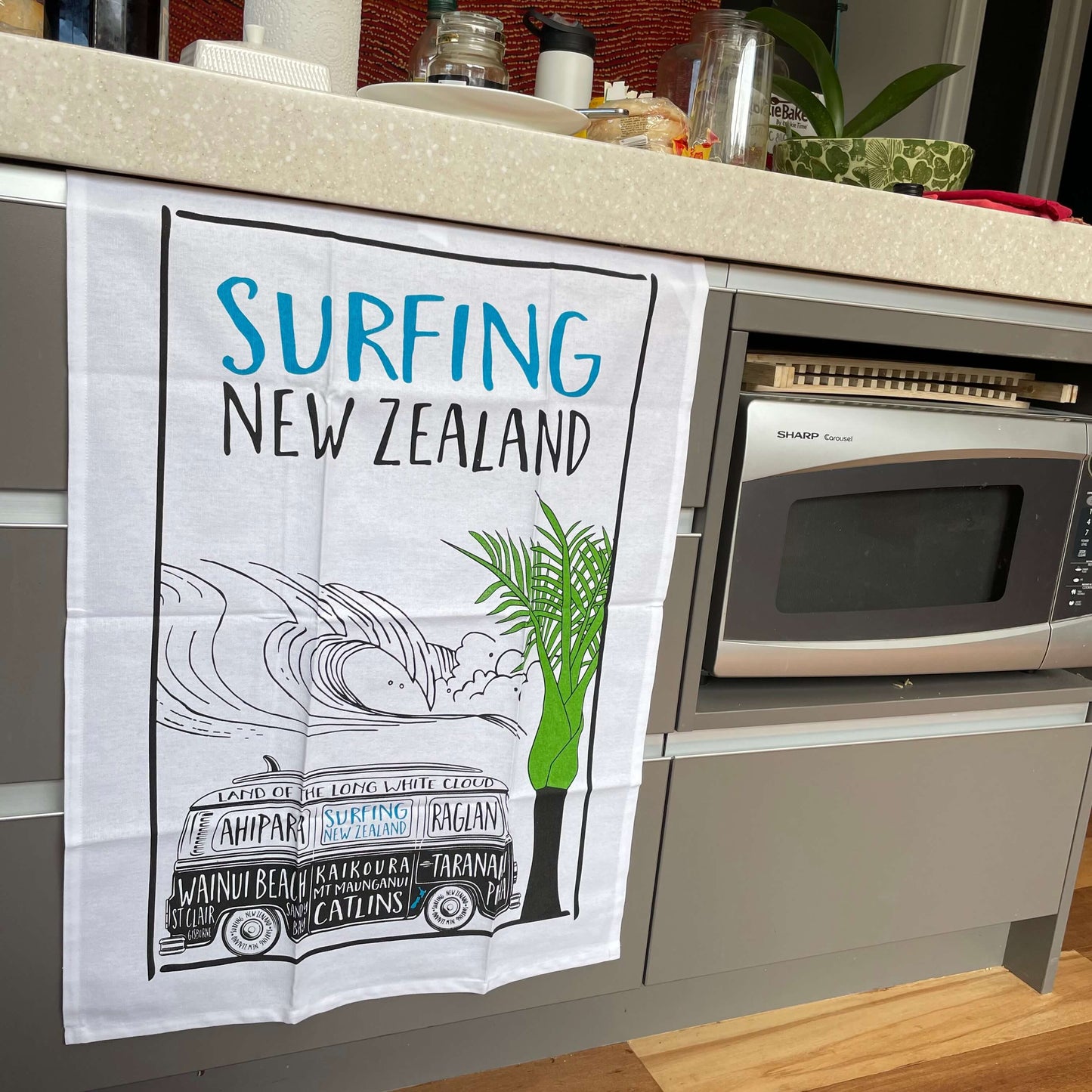 Surfing NZ tea towel with an image of a Kombi Van and NZ surf spots written on the van plus a green palm tree. Teatowel is hanging in front of kitchen drawers with items on the bench above and a microwave to the right.