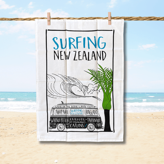 Surfing NZ tea towel with an image of a Kombi Van and NZ surf spots written on the van plus a green palm tree. Teatowel is hanging on a rope with 2 wooden pegs with a faded beach scene in the background.