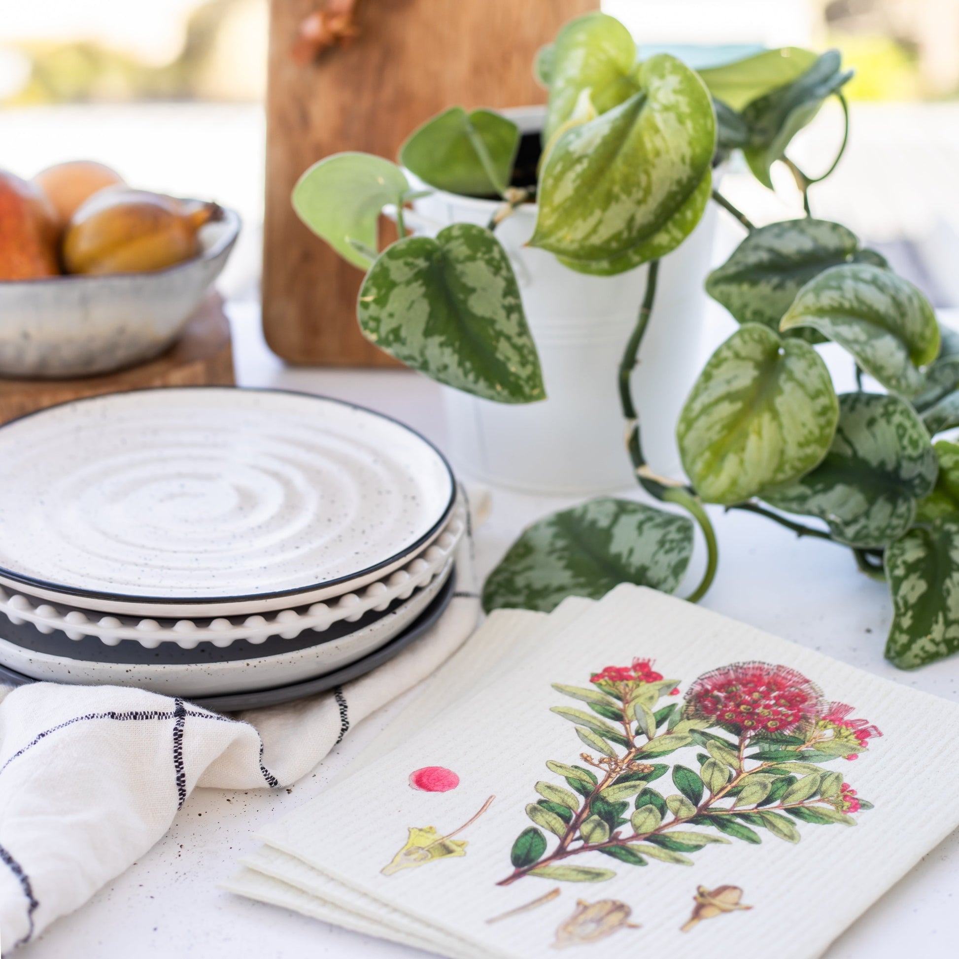 Set of 3 swedish dishcloths in New Zealand botanical prints on a table with plates, plants and a bowl of fruit.