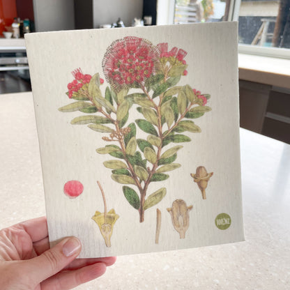 Hand holding a Swedish dishcloth with a kitchen in the background, The dishcloth has a picture of a New Zealand Northern Rata flower.