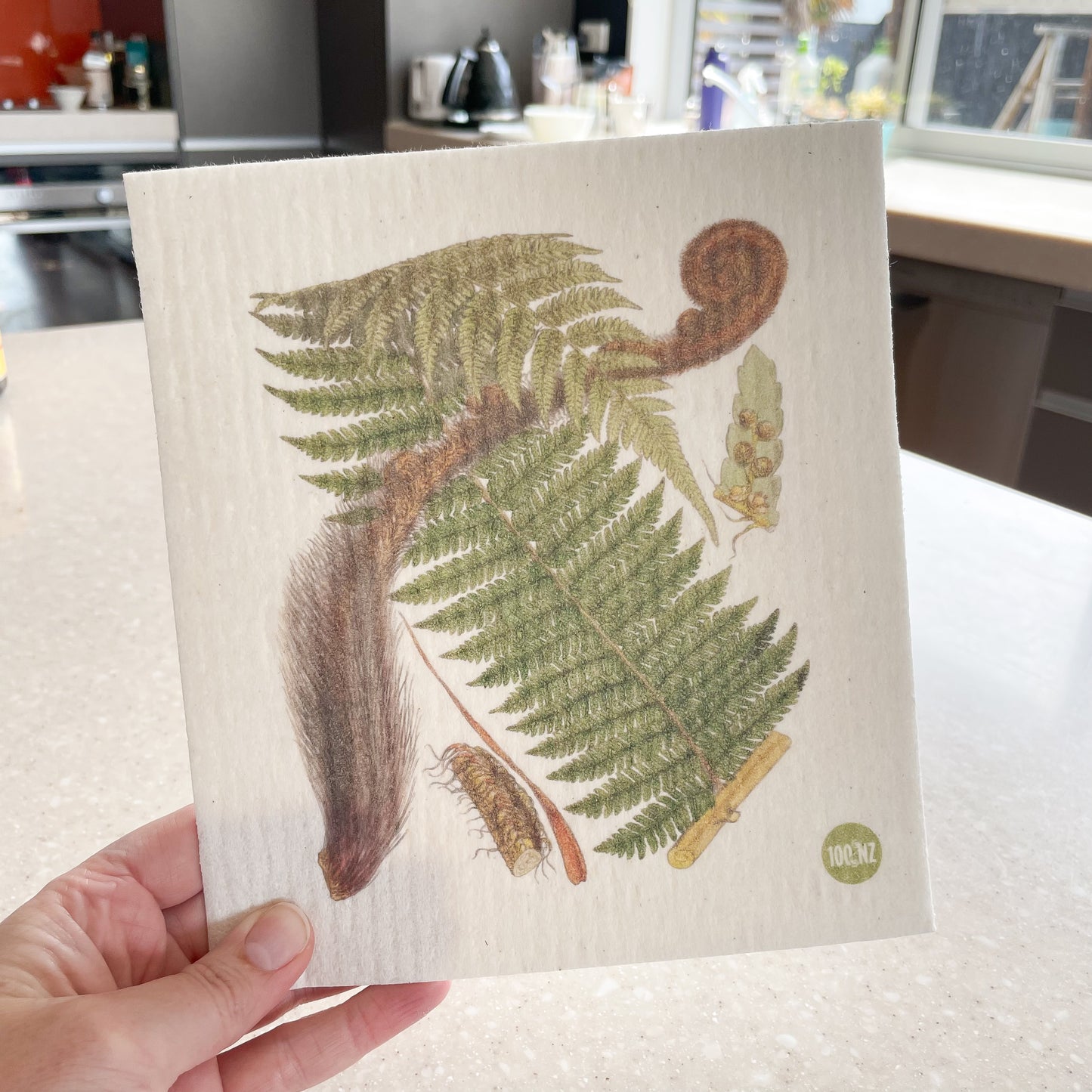 Hand holding a Swedish dishcloth with a kitchen in the background, The dishcloth has a picture of a native New Zealand Tree Fern on it.