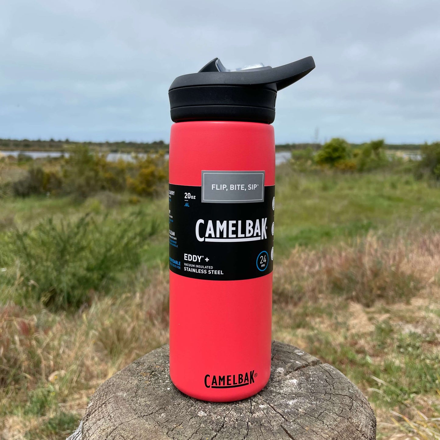 Stainless steel camelbak sipper bottle in Wild Strawberry colour with black lid. Sitting on a wooden fence post with a blurry Estuary scene in the background.