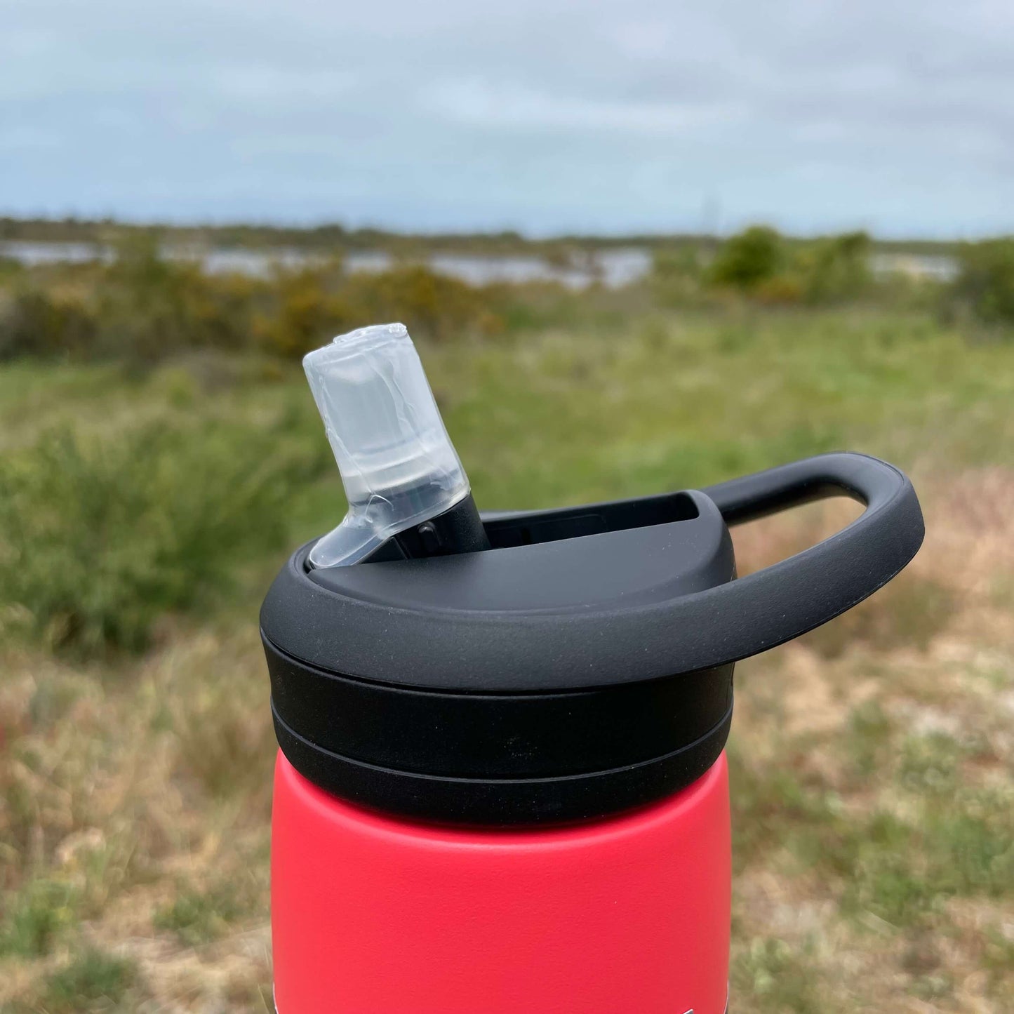 Close up of a black Camelbak lid with sipper top on a red drink bottle. Blurred out estuary in the background. 