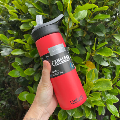 Persons hand holding a 600ml red stainless camelbak drink bottle with sipper open in front of a green hedge.