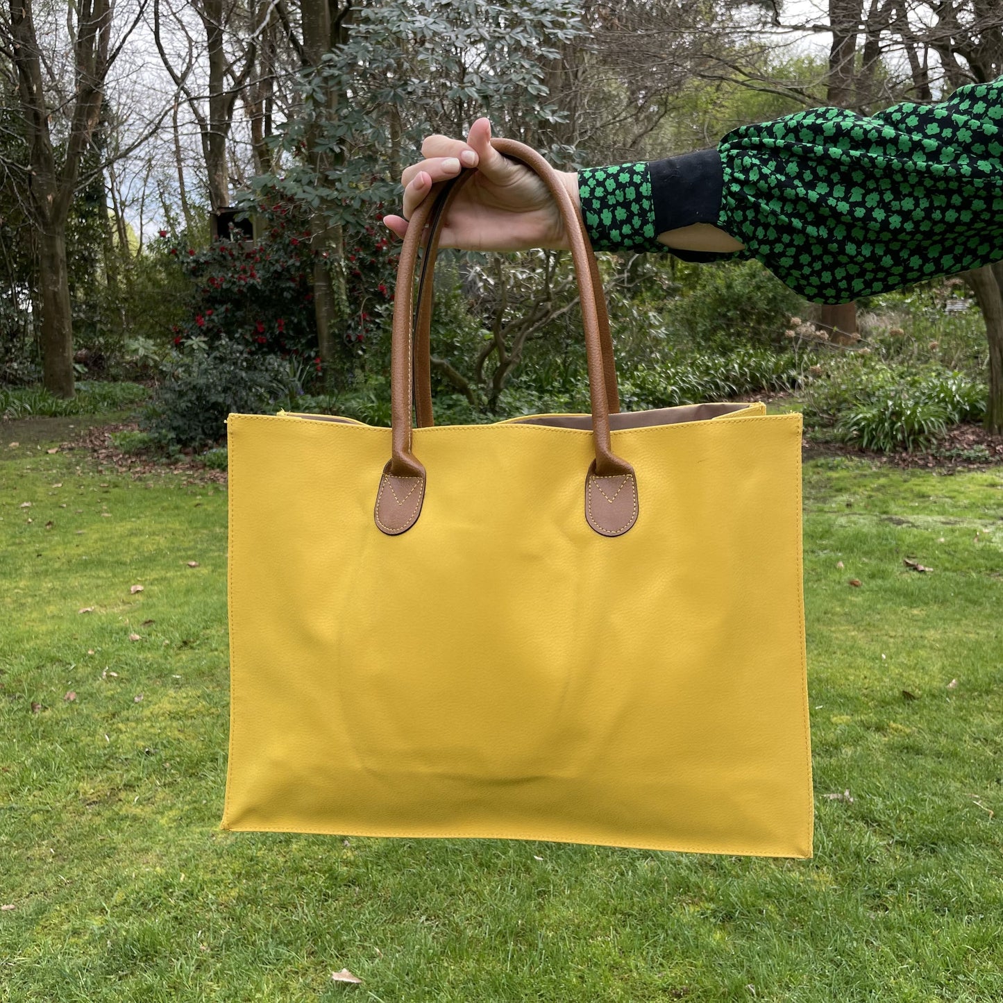 Womans hand holding a bright yellow tote bag with brown leather look handles in a garden setting.