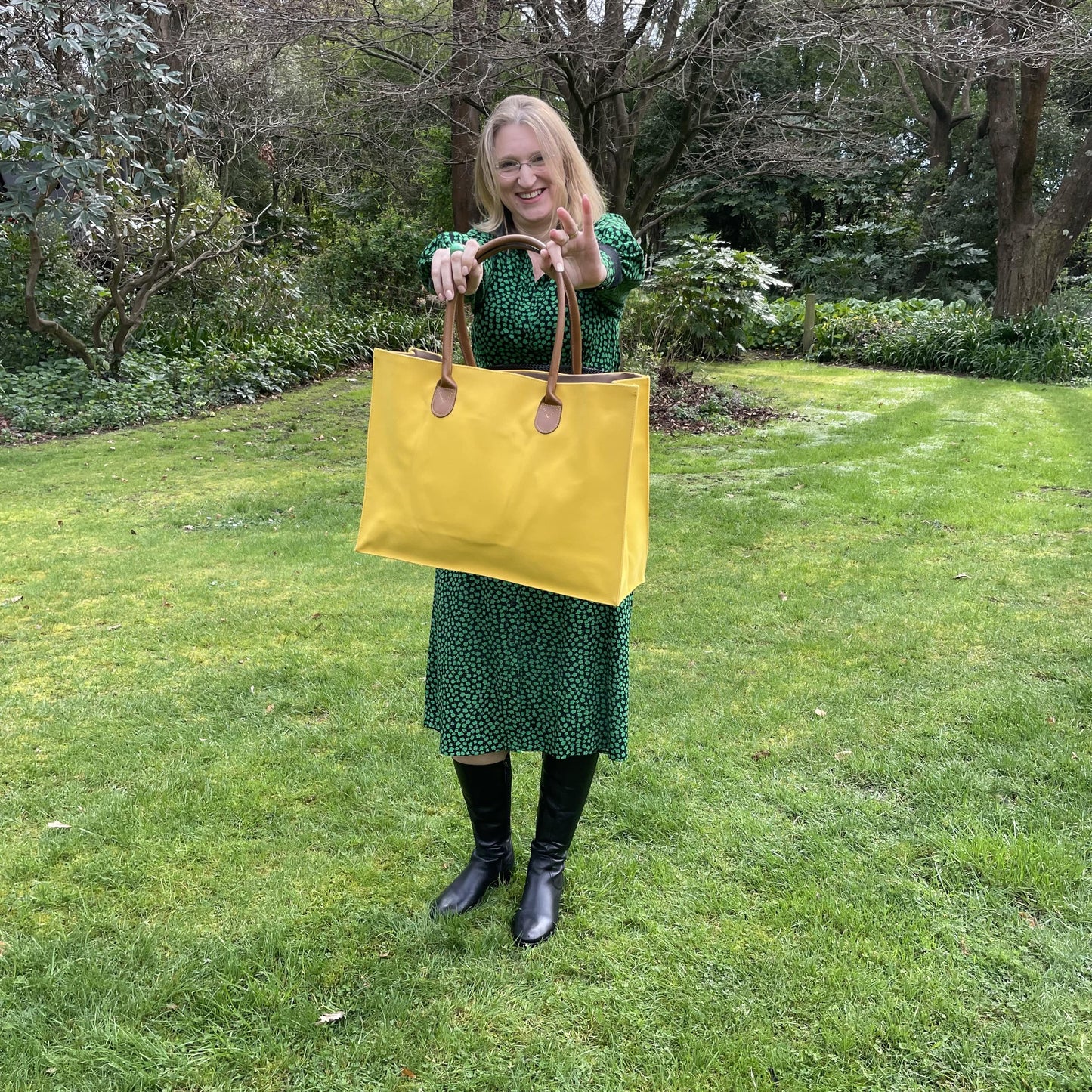 Woman wearing a green and black dress standing outside in a garden holding a large bright yellow tote bag by its brown handles out in front of her.