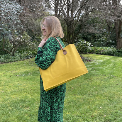 Woman with blonde hair and a green and black dress standing side on in a garden with a bright yellow tote bag over her shoulder.