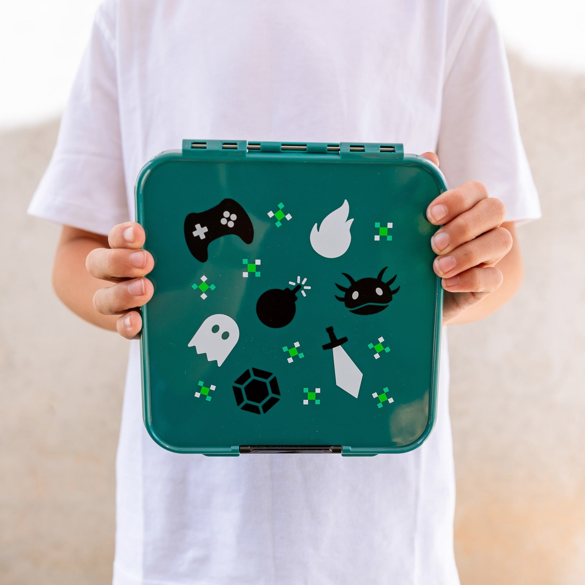 Close up of a childs hands holding a teal green lunchbox with a black and white retro gamer print on the lid.