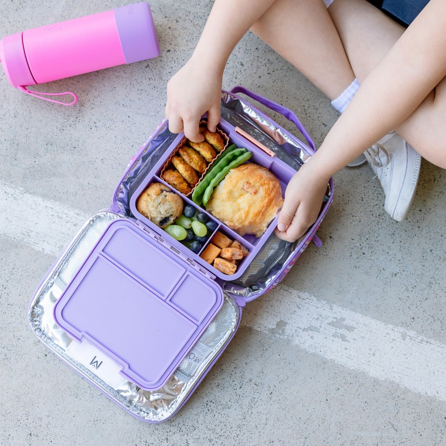 Child sitting crossed legged on the floor reaching into a bento style lunchbox filled with food.