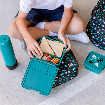 Young child sitting cross legged reaching for a sandwich from a teal green bento style lunch box and surrounded by matching acessories; drink bottle, snack box and backpack.