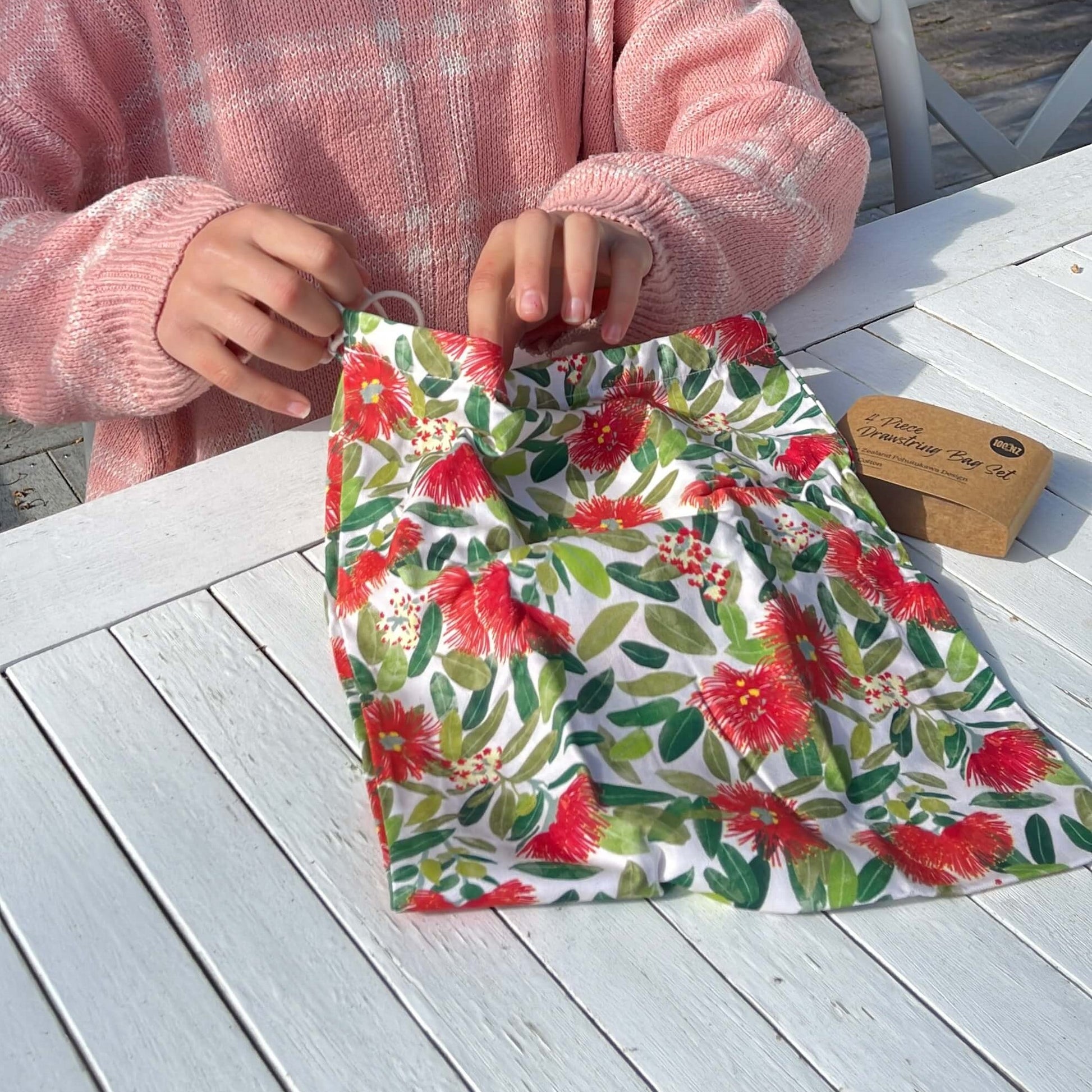 Womans hands opening a pohutukawa print drawstring bag.