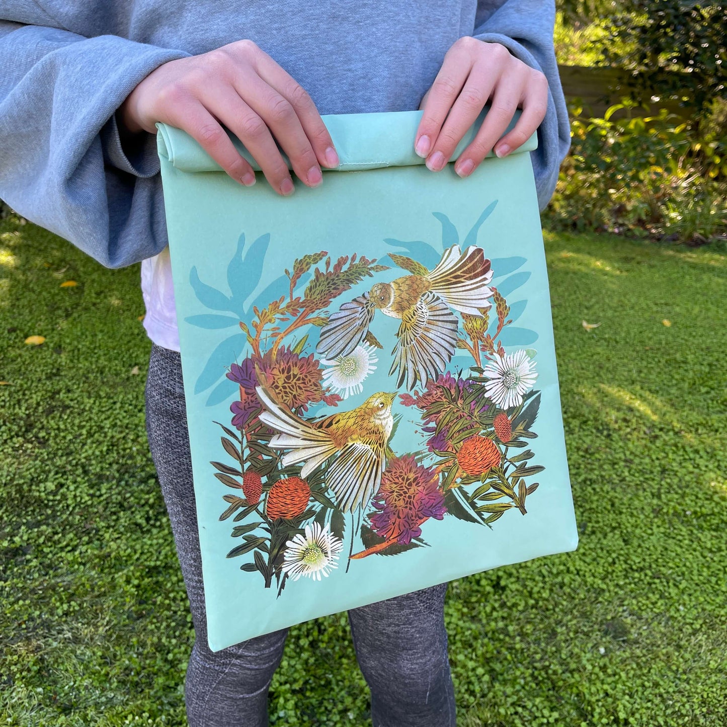 Person holding a light blue folding paper style lunch bag with floral and bird design.