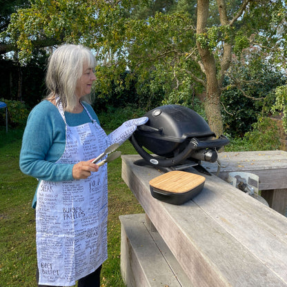 Woman wearing a white apron and mitt set featuring recipes printed on it standing outside at a BBQ.