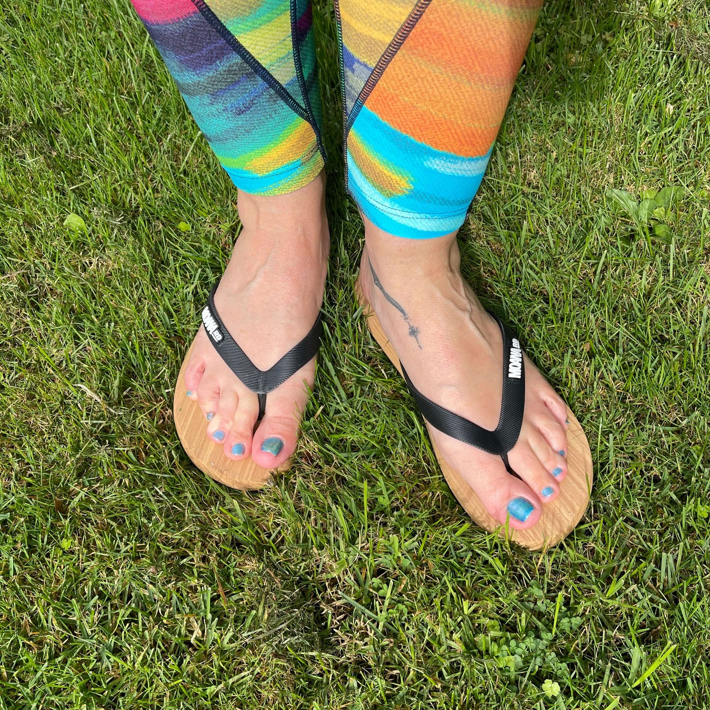 Womens feet wearing unisex Jandals with a wood grain look and black straps standing on grass.