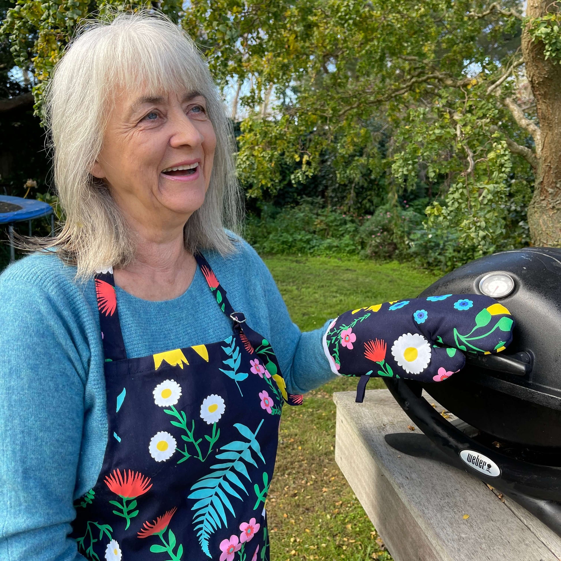 Woman wearing a navy blue and floral apron and mitt set standing outside at a BBQ.