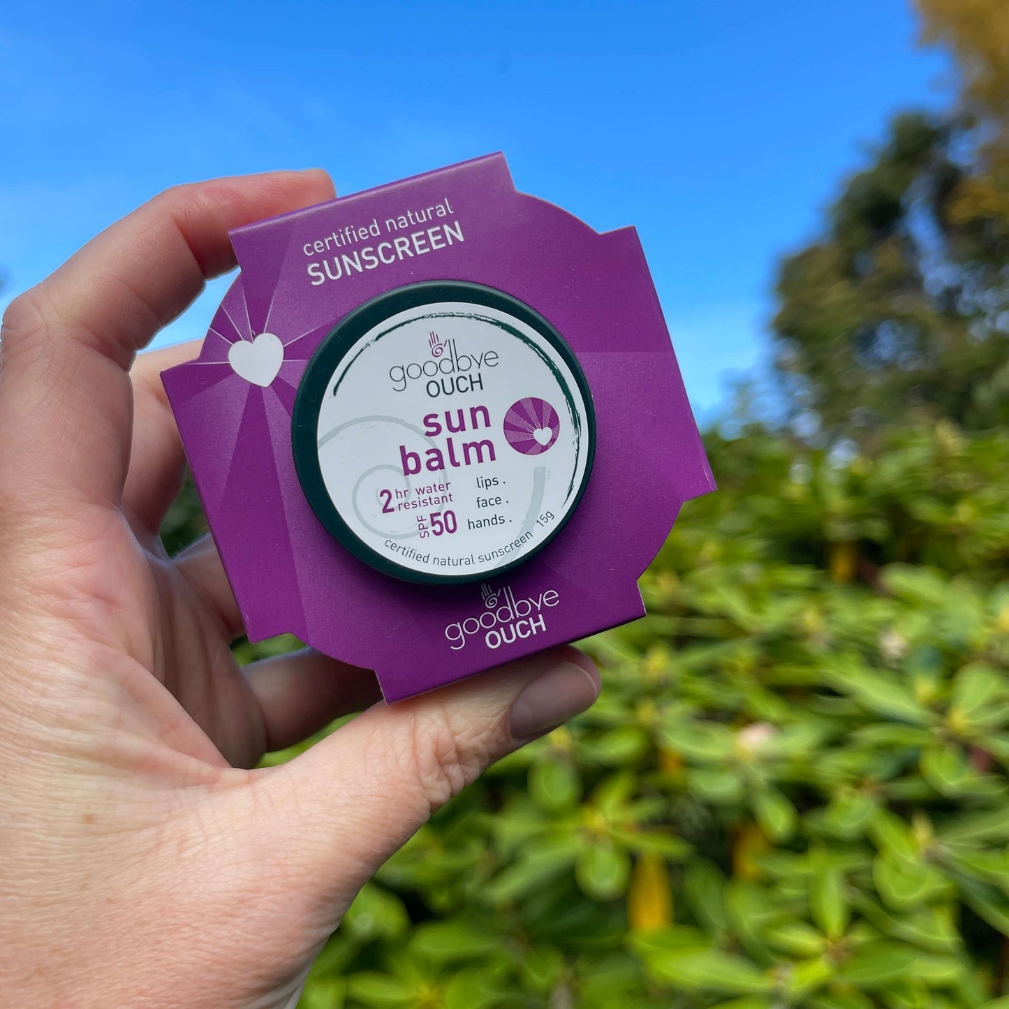 Womens hand holding a sun balm starter kit from Goodbye Ouch with blue sky in the background.